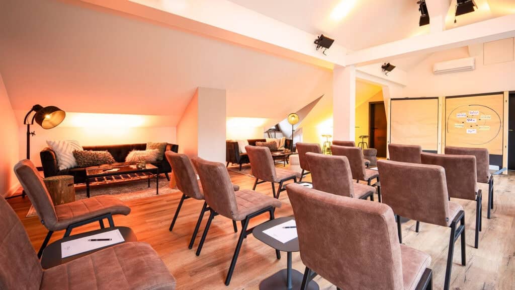 Ein moderner Seminarraum am Campus 54:2 mit Reihen brauner Stühle für Führungskräfte Seminare und Young Professionals, einer Leinwand, Sofas im hinteren Bereich, warmer Beleuchtung und Notizblöcken auf jedem Platz.