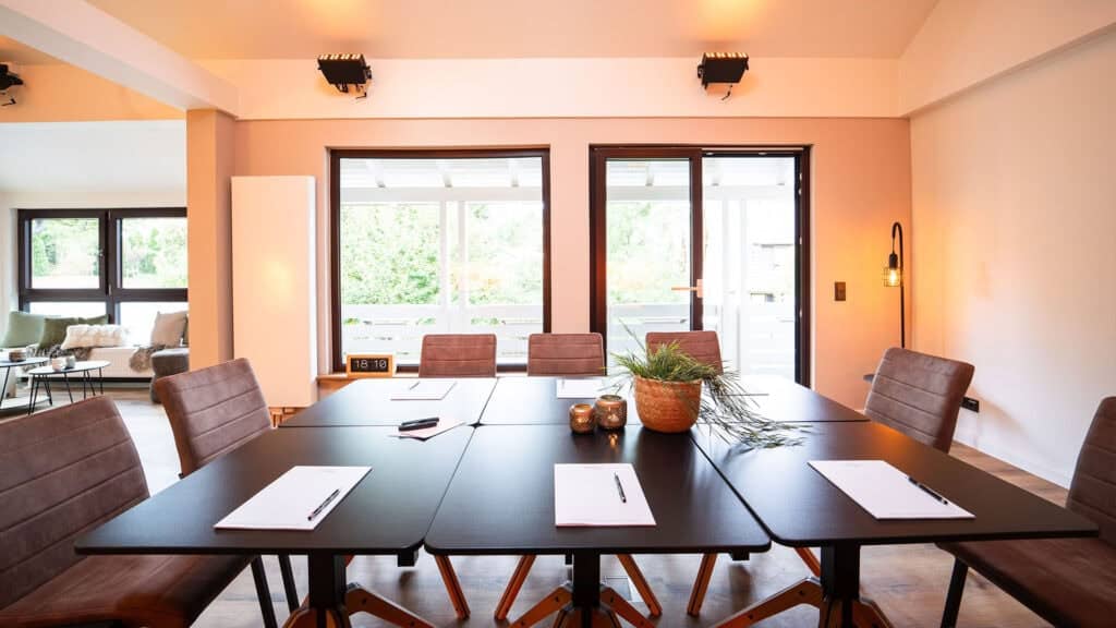 Ein moderner Konferenzraum am Campus 54:2 mit einem schwarzen Tisch, braunen Stühlen, Notizblöcken, Stiften, einer Pflanze und Kerzen – ideal für Führungssitzungen oder Führungskräfteseminare – in der Nähe großer Fenster und Glastüren, die natürliches Licht hereinlassen.