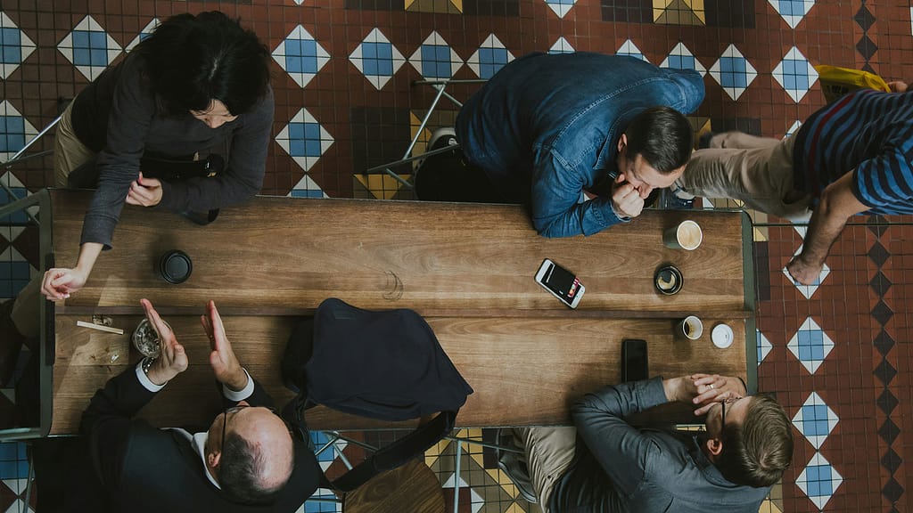 Vier junge Berufstätige versammeln sich mit Getränken und einem Telefon um einen Holztisch. Die Ansicht ist von oben zu sehen, während eine fünfte Person auf dem gemusterten Fliesenboden vorbeigeht – ein Eindruck vom kooperativen Geist, der in Führungs- und Führungskräfteseminaren herrscht.