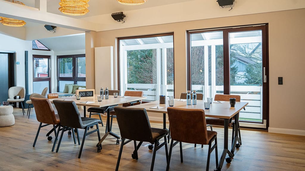 Ein Konferenzraum mit braunen Stühlen, Tischen, die für ein Führungstreffen aufgestellt wurden, Wasserflaschen, Notizblöcken und einem Timer auf dem Tisch, mit großen Fenstern, die für natürliches Licht sorgen.