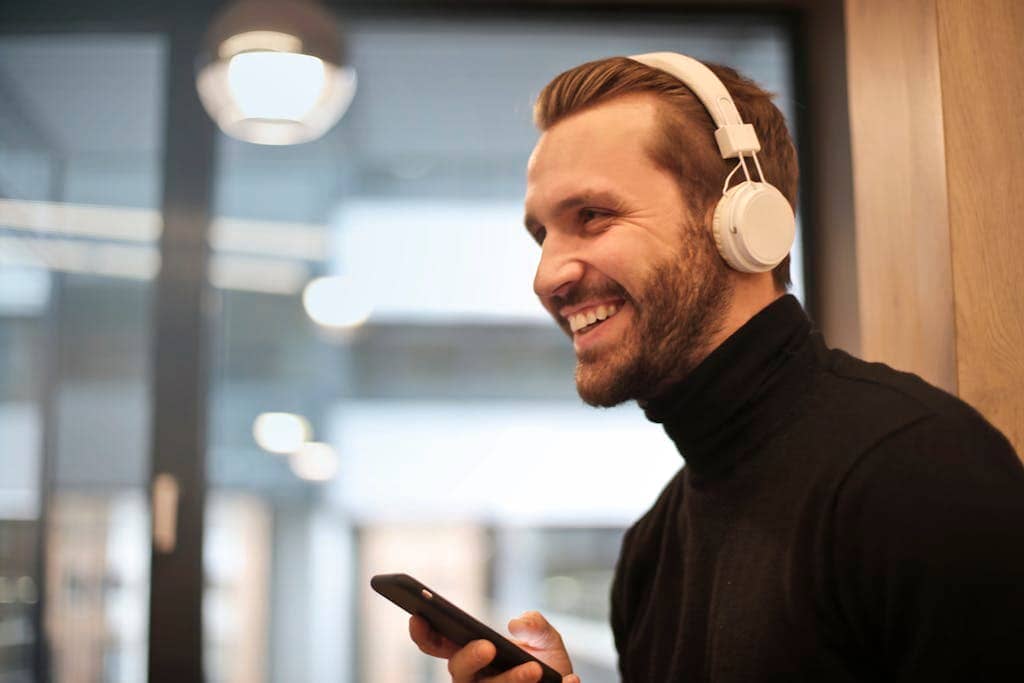 Ein Mann mit Kopfhörern und schwarzem Rollkragenpullover lächelt, während er drinnen ein Smartphone hält und den selbstbewussten Geist junger Berufstätiger verkörpert.