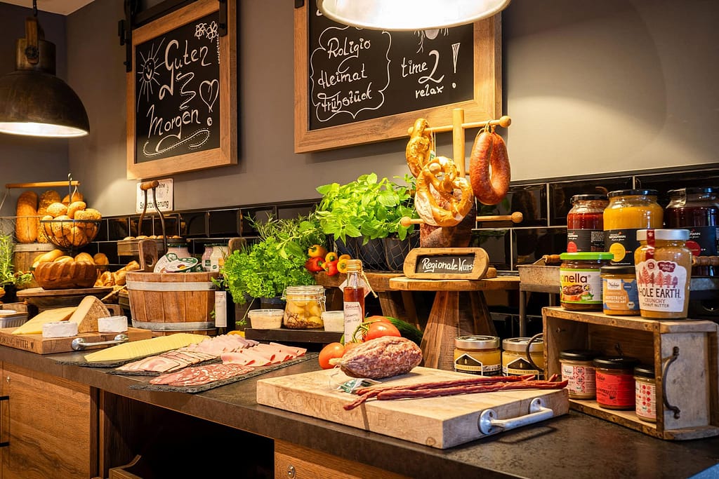 Ein Frühstücksbuffet mit verschiedenen Brotsorten, Aufschnitt, Wurst, Käse, Aufstrichen, frischem Gemüse und Gläsern mit Gewürzen, angeordnet auf einer Theke unter Tafelschildern – der perfekte Energielieferant für junge Berufstätige und Führungskräfte, um in den Tag zu starten.