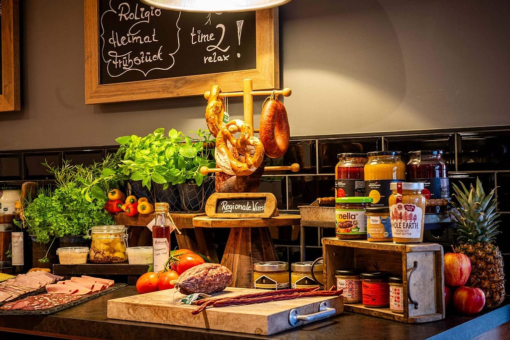 Ein Frühstücksbuffet für junge Berufstätige umfasst verschiedene Fleischsorten, Brot, frische Kräuter, Obst und Gläser mit Aufstrichen, kunstvoll arrangiert auf einer schwarzen Theke unter einem Schild aus Kreidetafel.