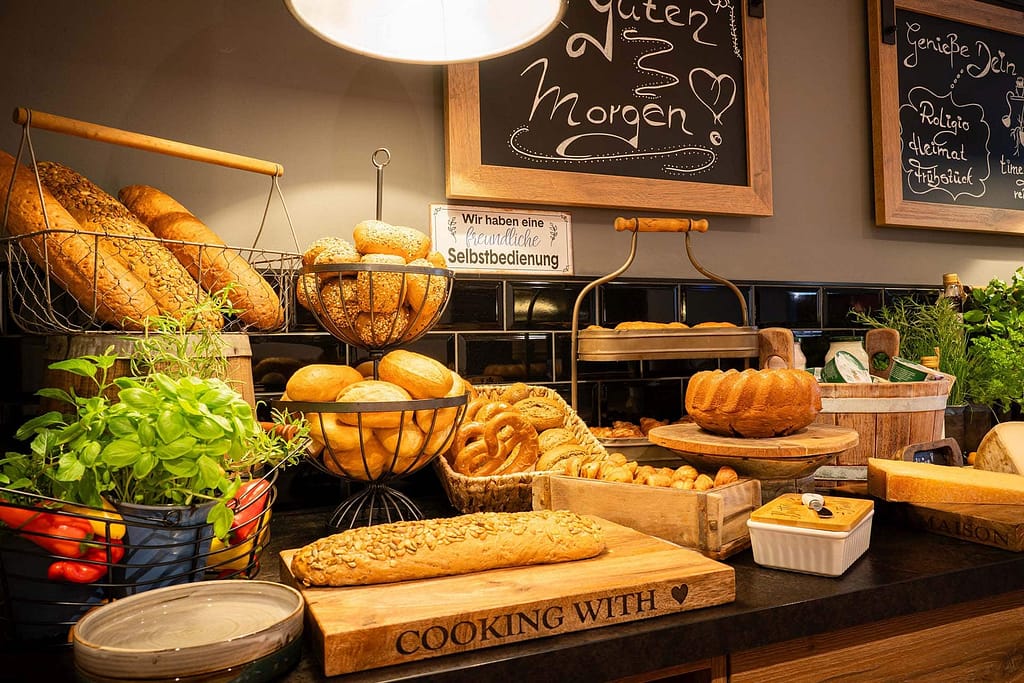 Eine Auswahl an Brot und Gebäck wird auf einer Holztheke präsentiert, im Hintergrund sind Körbe, Schneidebretter, Kräuter und Kreidetafelschilder zu sehen – perfekt für junge Berufstätige, die Wert auf Geschmack und Stil legen.