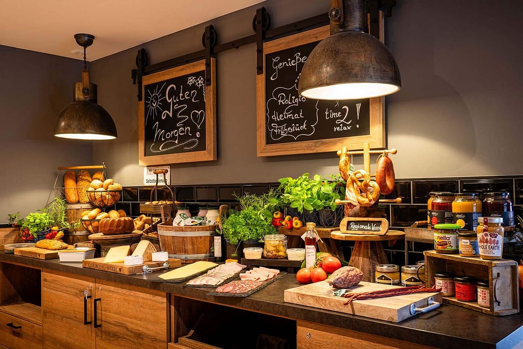 Unter Tafelmenüs und Industriebeleuchtung erwartet Sie ein Frühstücksbuffet mit Brot, Aufschnitt, Käse, Aufstrichen, Gemüse, frischen Kräutern und hängenden Wurstwaren – perfekt für Gäste der Generation Gold oder Campus 54:2 Führungskräfte-Seminare.