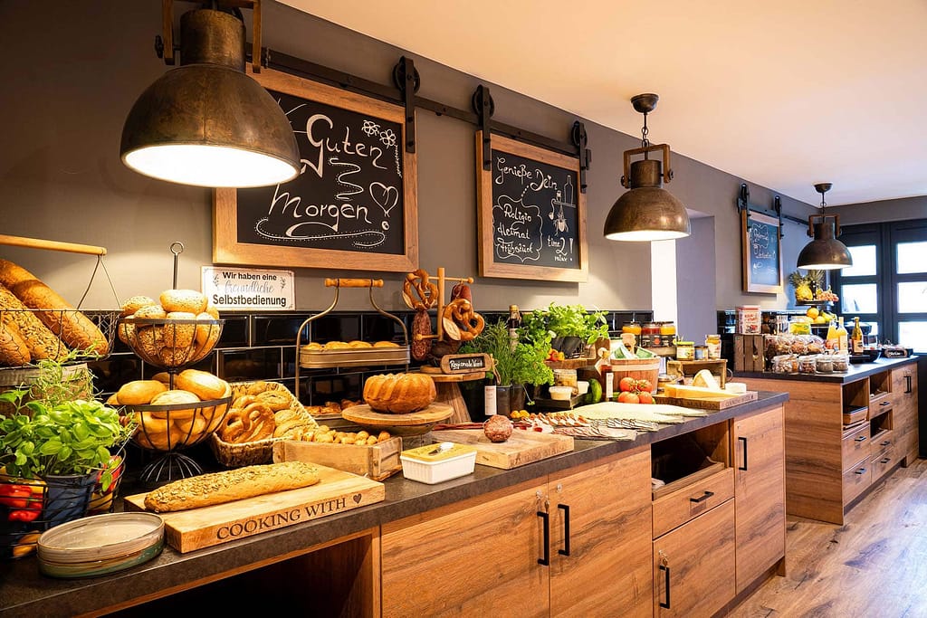 Genießen Sie im Campus 54:2 ein Frühstücksbuffet mit Brot, Gebäck, Aufschnitt, Käse, Obst und Kräutern – ein perfekter Start vor Ihrem Führungskräfteseminar in einem gut beleuchteten, mit Tafeln geschmückten Raum.