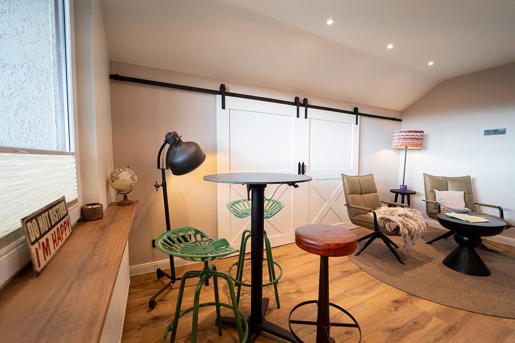 Ein moderner Raum für junge Berufstätige mit einem hohen Tisch und Hockern, Sesseln, einer Stehlampe, einer Schiebetür im Scheunenstil und einer Fensterbank mit stilvollen Dekorationsgegenständen.