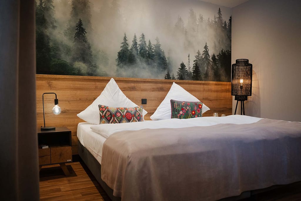 Ein ordentlich gemachtes Doppelbett mit weißer Bettwäsche, gemusterten Kissen, einem hölzernen Kopfteil und einem Wandgemälde eines nebligen Waldes; Nachttische mit Lampen auf beiden Seiten schaffen einen beruhigenden Rückzugsort, inspiriert vom Stil von Campus 54:2.