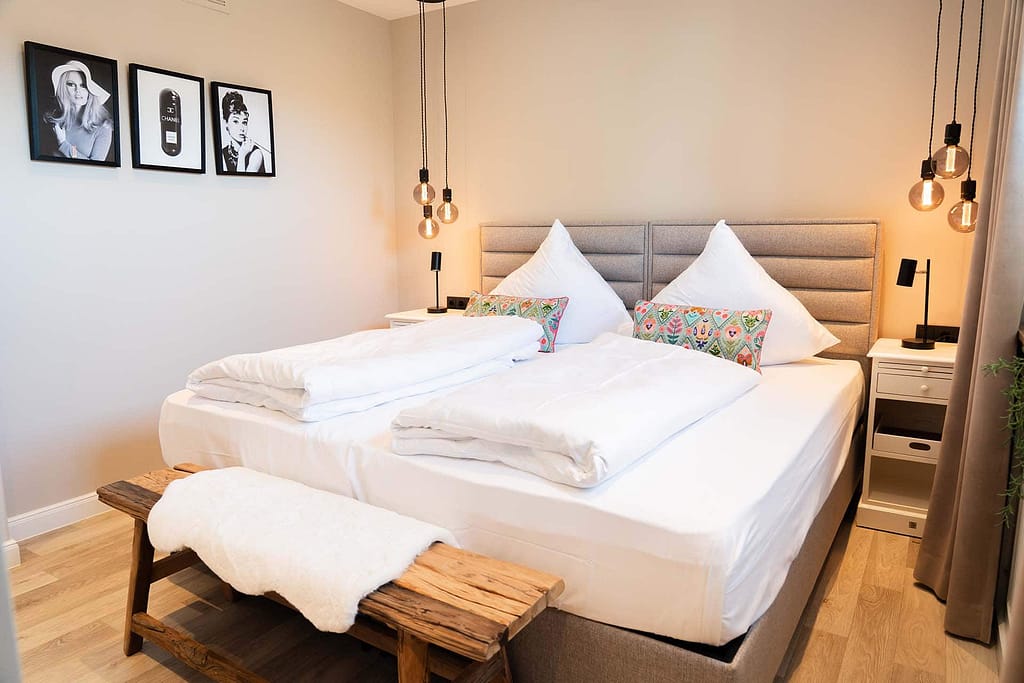 Ein ordentlich gemachtes Doppelbett mit weißer Bettwäsche, zwei bunten Kissen und einer Holzbank in einem modernen Schlafzimmer – perfekt für junge Berufstätige im Campus 54:2, mit Hängelampen und gerahmten Fotos, die stilvolle Akzente setzen.