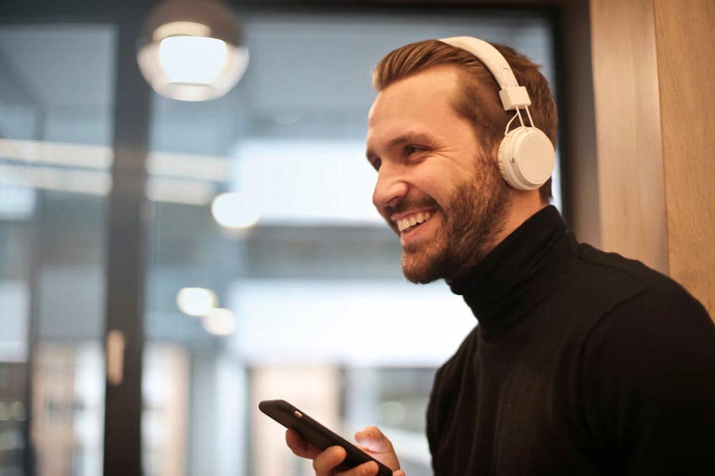 Ein Mann mit Kopfhörern und schwarzem Rollkragenpullover lächelt, während er drinnen ein Smartphone hält und den selbstbewussten Geist junger Berufstätiger verkörpert.