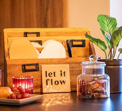 Ein Organizer aus Holz hält Umschläge neben einem Glas mit Trockenfrüchten, einer Topfpflanze, Kerzen, Weintrauben, Brot und einem „Let it flow“-Schild auf dem Tisch – perfekt für inspirierende Momente beim Campus 54:2 oder den Generation Gold Führungskräfte Seminaren.