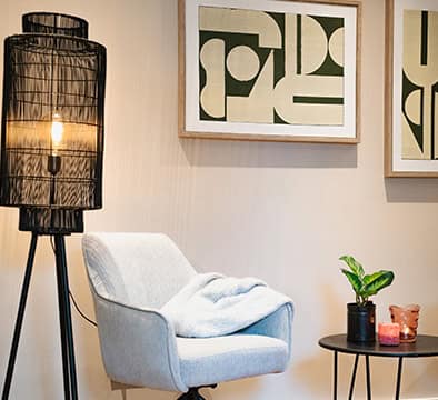 Eine moderne Wohnzimmerecke für junge Berufstätige mit einem grauen Sessel, einer Stehlampe, einem Beistelltisch mit einer Pflanze und Kerzen sowie zwei abstrakten Kunstwerken an der Wand.