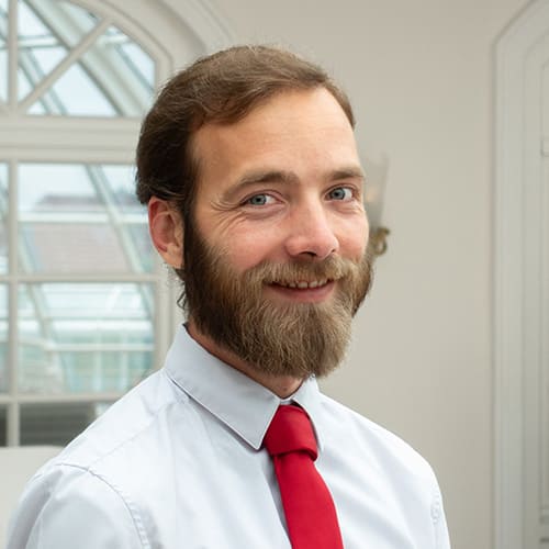 Mann mit Bart und Schnurrbart, weißes Hemd und rote Krawatte, steht in einem Innenraum vor einem großen Bogenfenster.