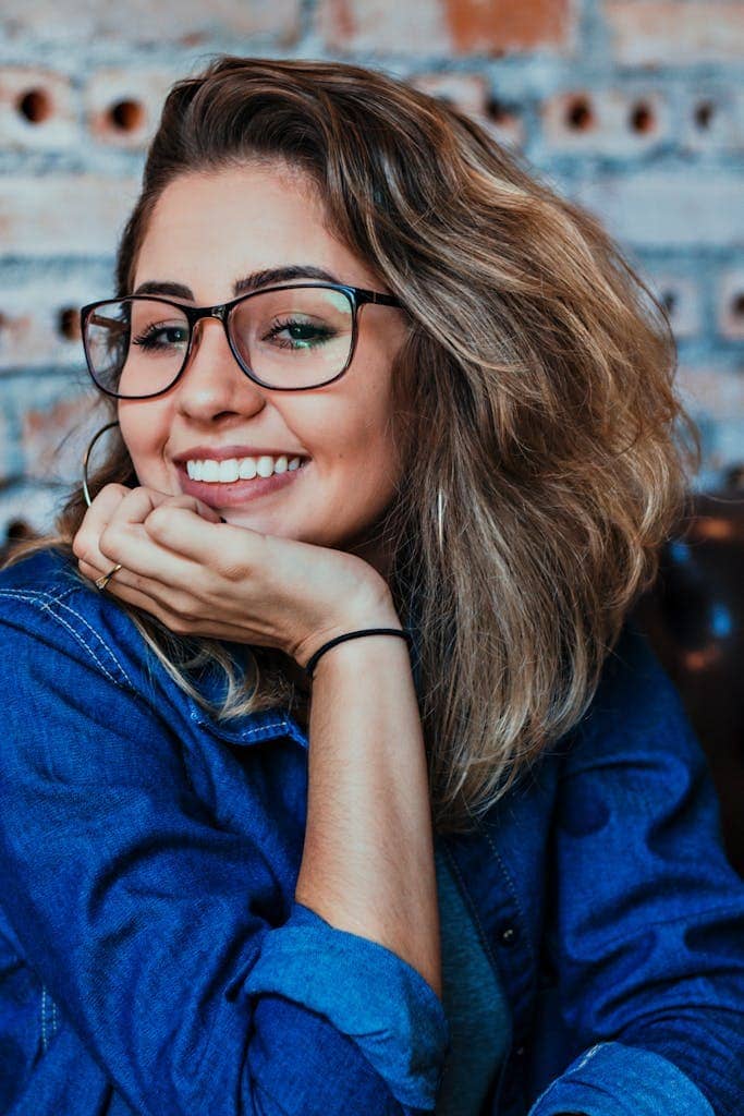 Eine Frau mit welligem Haar und Brille, die ein Jeanshemd trägt, lächelt, während sie vor dem Hintergrund einer Backsteinmauer ihr Kinn auf ihre Hand stützt – und strahlt damit Selbstvertrauen und Führungsstärke aus.