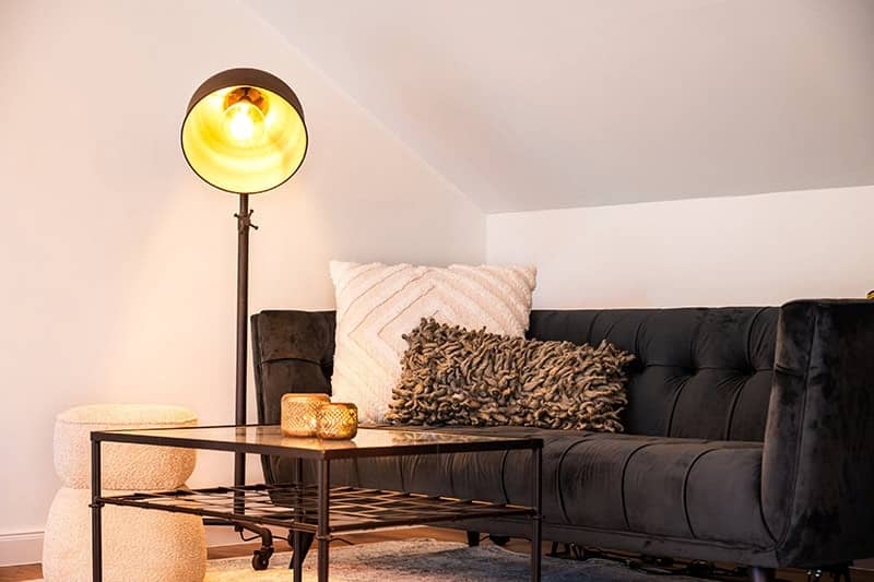 Ein modernes Wohnzimmer für junge Berufstätige mit einem schwarzen Sofa, strukturierten Kissen, einer Stehlampe mit warmem Licht, einem schwarzen Couchtisch und zwei runden Sitzkissen.